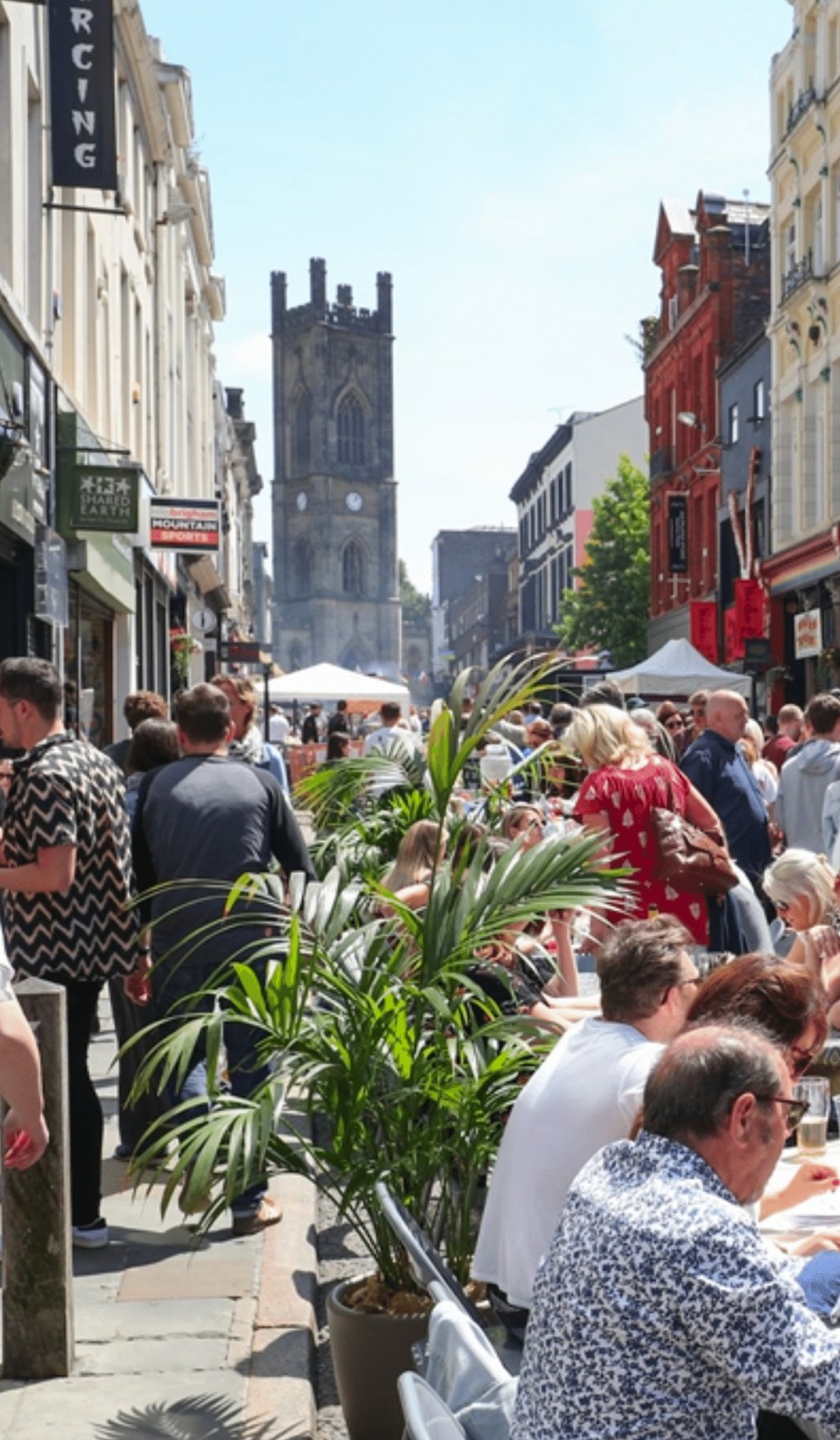 A very busy bold street on a clear sky day in liverpool, with a flock of people walking down the narrow street and other people having lunch at the restaurants and cafes