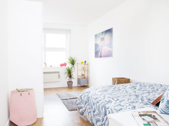 Big modern bedroom with wooden laminate floors. The bedroom is fully furnished with a queen bed, a grey rug, a pink chequered laundry basket, and a small closet. The Room is decorated with one square window, a tall plant, a holographic poster, a floor cupboard. 